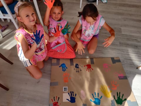 Three young children in pink clothes displaying handprints and finger paintings on a large cardboard surface during an art activity