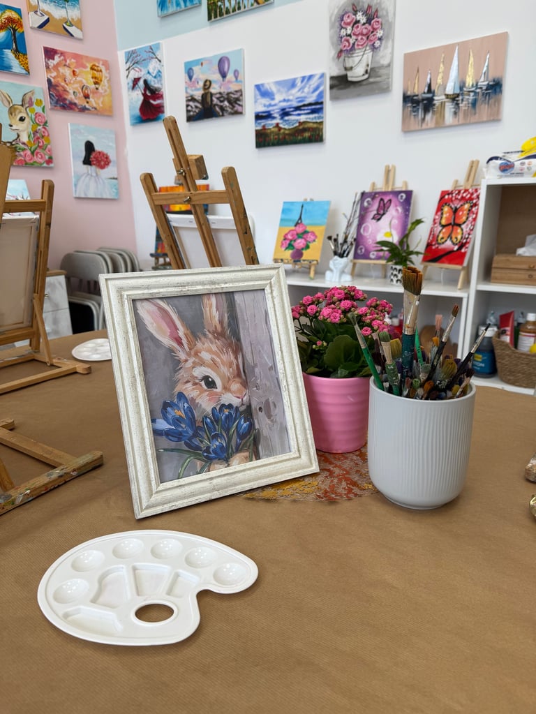 Artist studio workspace with framed watercolor rabbit painting, paint palettes, brushes in cups, easels, and artwork displayed on walls