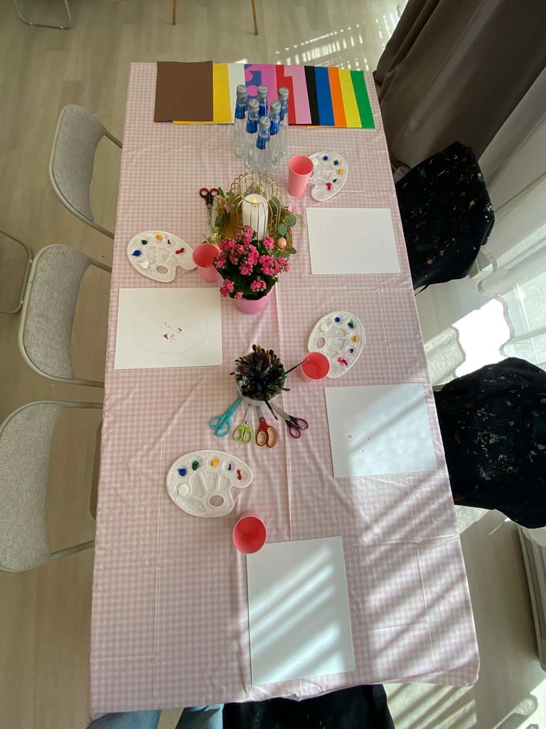 Art class setup with pink checkered table, paint palettes, potted flowers, colored paper, and crafting supplies arranged for group activity