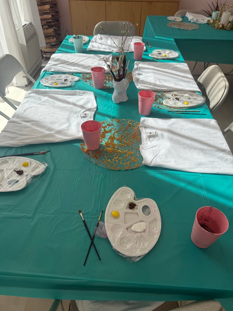 Art party table setup with turquoise tablecloth, white napkins, paint palettes, brushes in vases, and pink cups for an indoor painting activity
