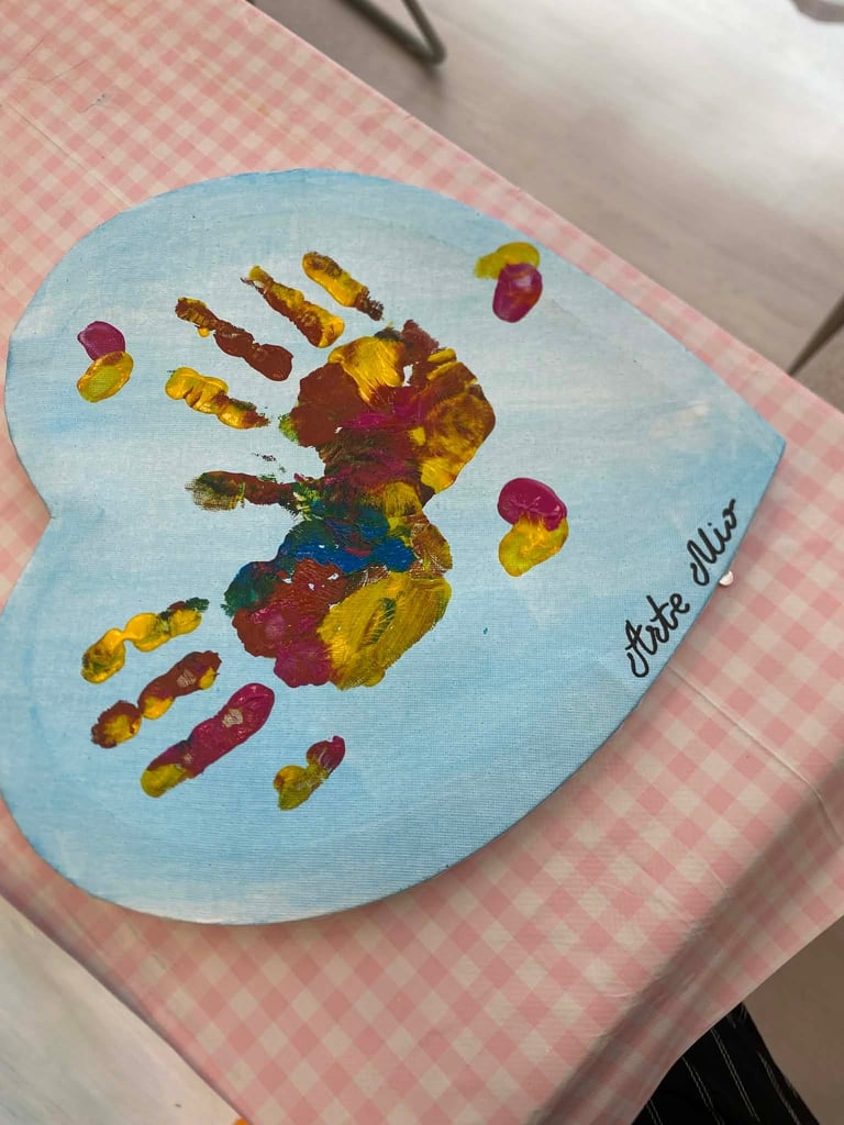 Childs handprint art on light blue heart-shaped paper with colorful paint in red, yellow, and green, with text reading I love you on pink checkered fabric