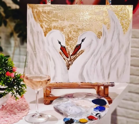 Canvas with white swan painting and gold leaf detail, surrounded by art supplies, wine glass, and flowers on wooden easel