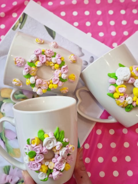 White ceramic mugs and dishes decorated with colorful clay flowers against a pink polka dot background