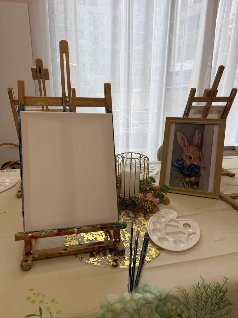 Wooden easel with blank canvas next to framed rabbit portrait, with painting supplies and palette on table