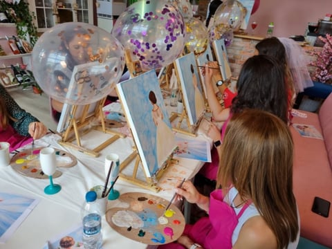 Two girls painting at an art table with easels, decorative balloons, and craft supplies in a colorful studio space
