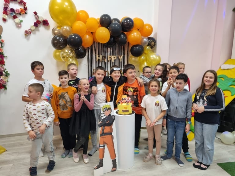 Group of children posing together under a yellow, orange, and black balloon arch decoration at an indoor party