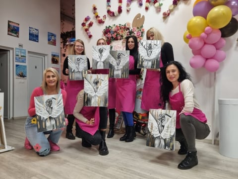 Group of women holding artwork at a gallery event decorated with pink, yellow, and black balloons and floral arrangements